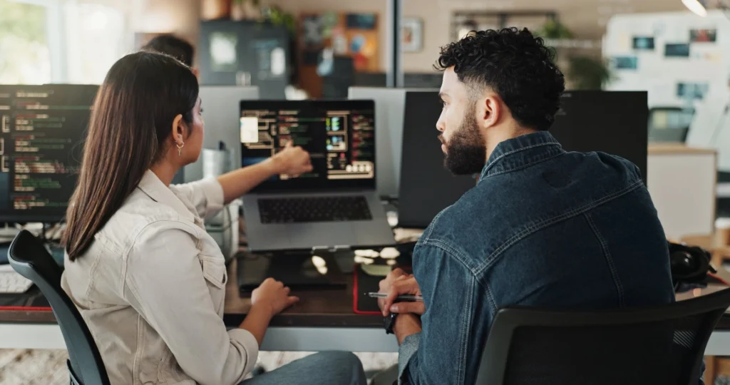 Two developers collaborating at desk while reviewing code on laptop